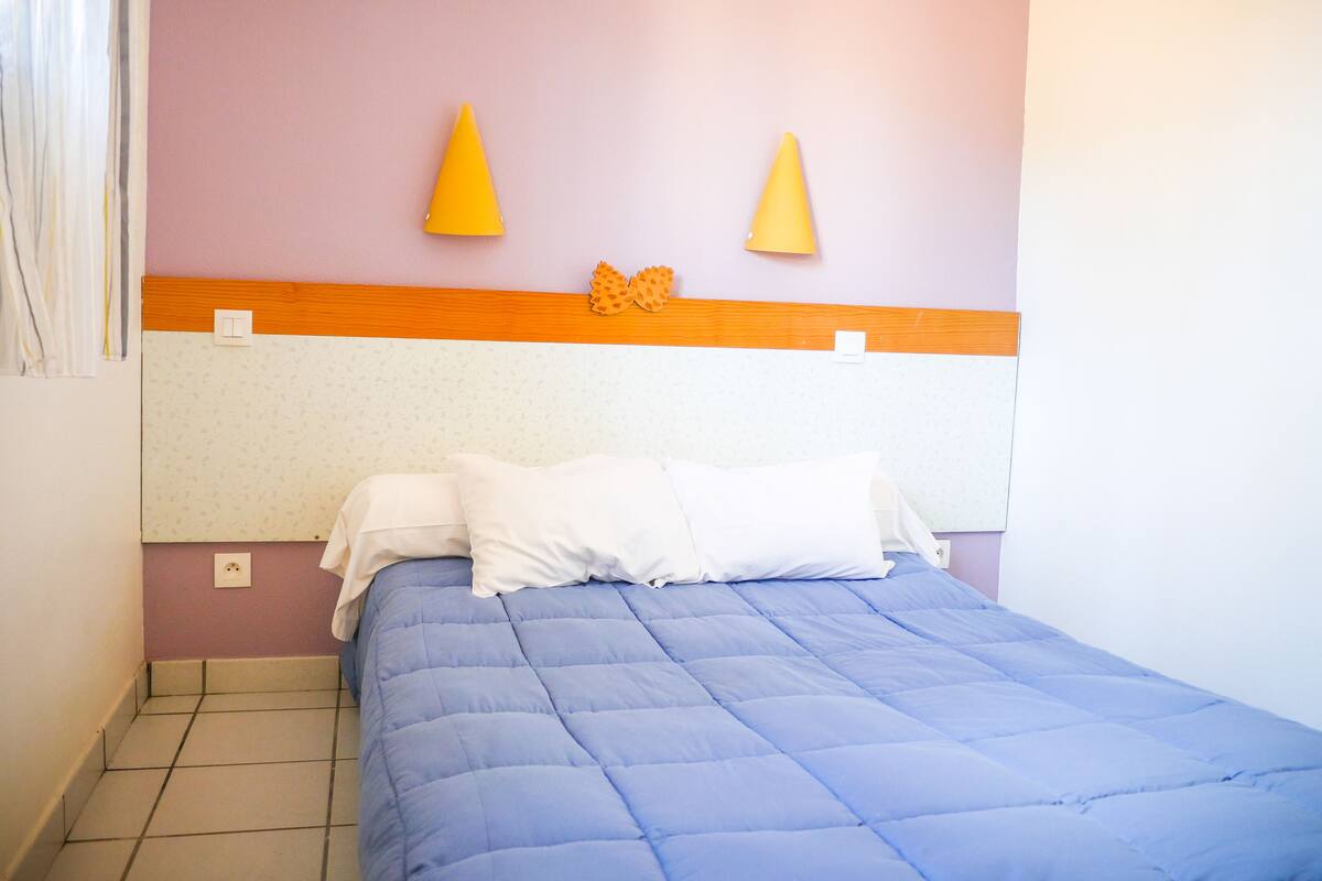 A bedroom with a bed, a headboard, a wall-mounted light, and a power outlet.