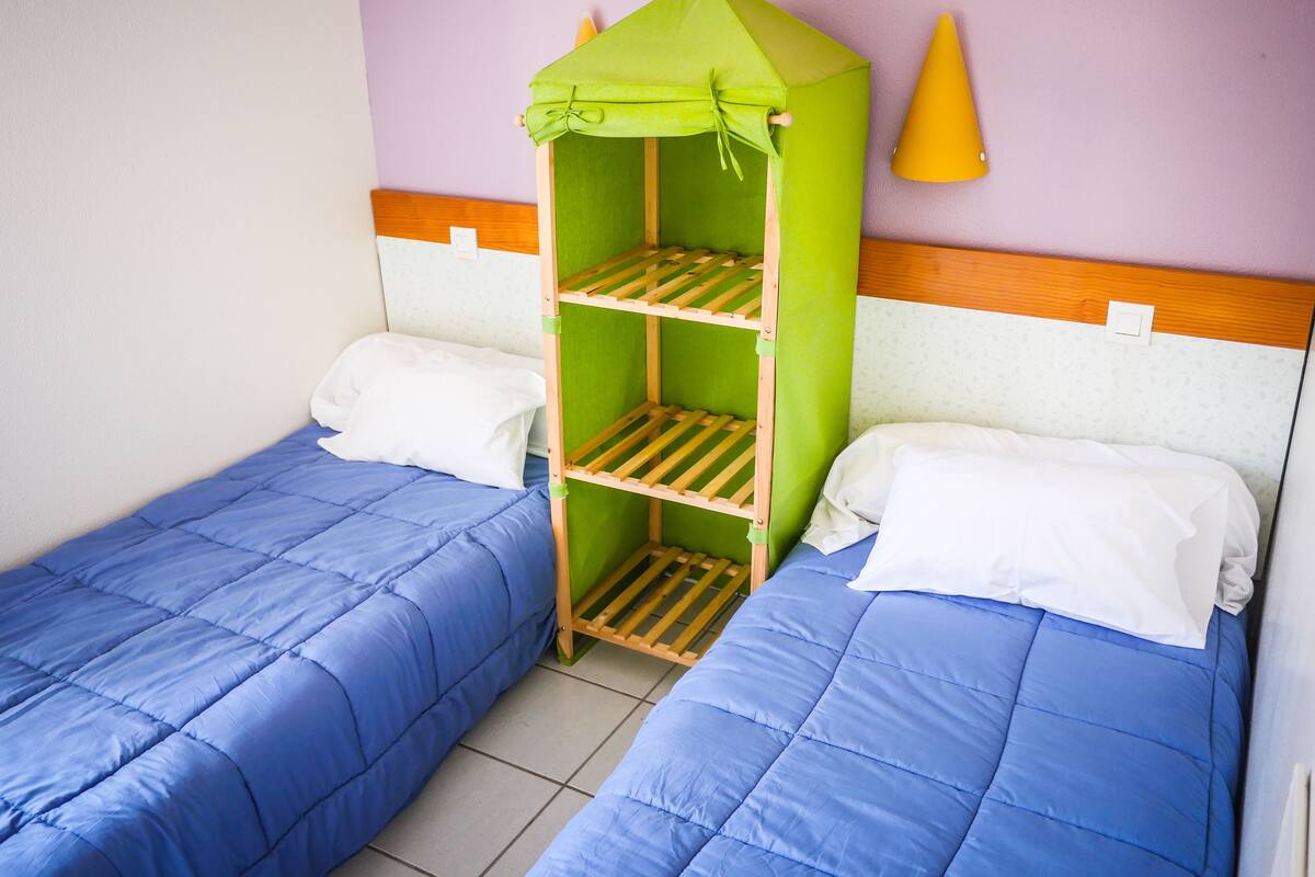 A room with two single beds, each with blue bedding and white pillows. A green, house-shaped bunk bed is positioned between the beds.