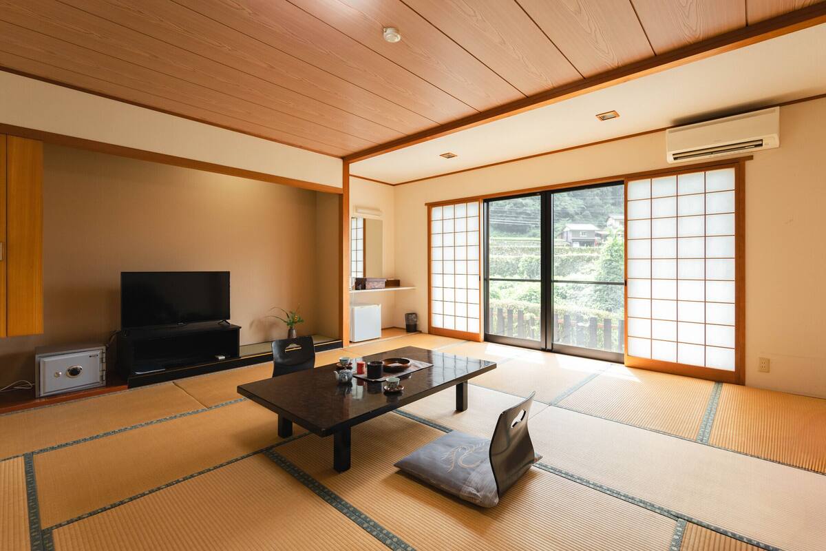 Une pièce de style japonais traditionnel, avec un sol recouvert de tatamis, une télévision, un petit meuble et une porte coulissante donnant sur un balcon.