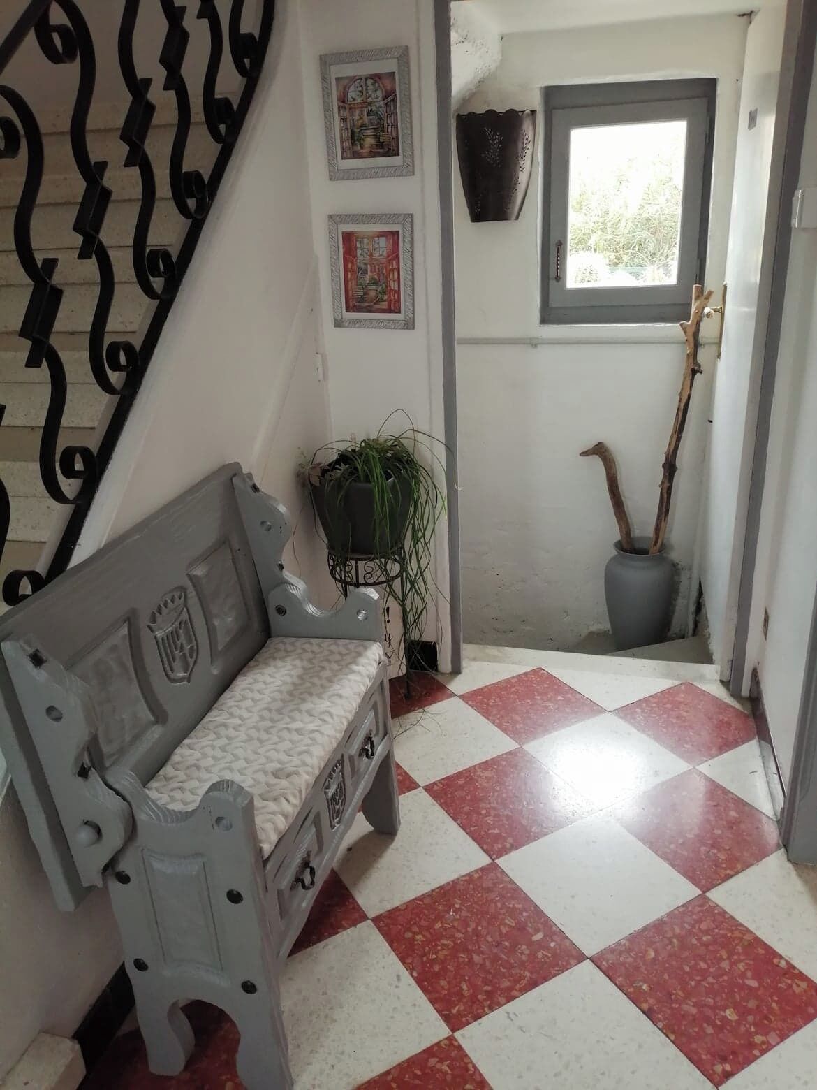 Une pièce avec un sol à carreaux, un banc, une plante en pot, une fenêtre et un élément décoratif fixé au mur.