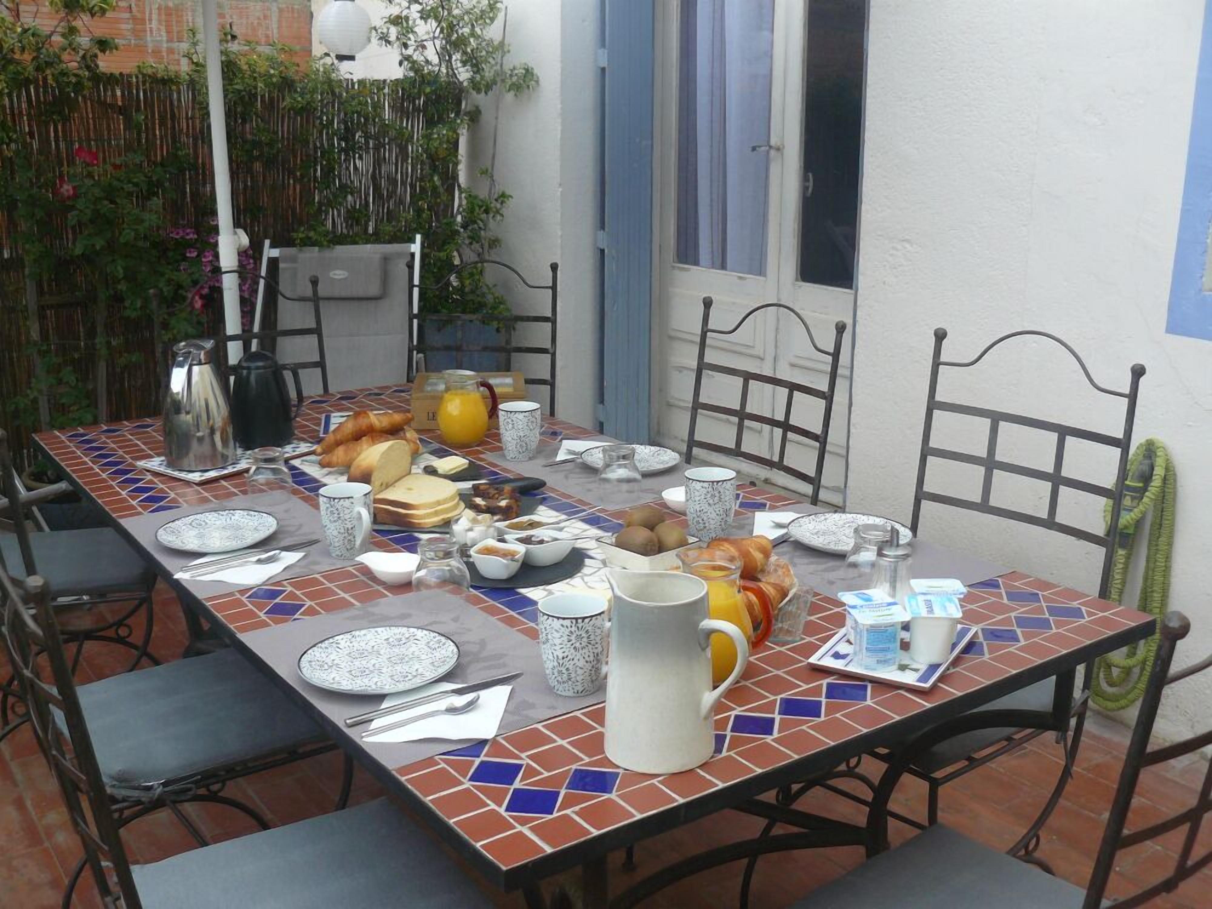 A dining table set with breakfast items, surrounded by chairs, in a courtyard with a bamboo fence and a white wall.