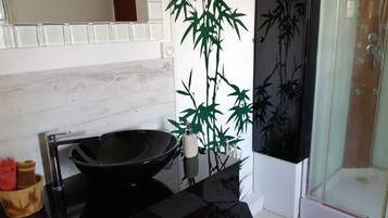 Une salle de bain avec un lavabo noir, un miroir, une plante en bambou et une cabine de douche en verre.