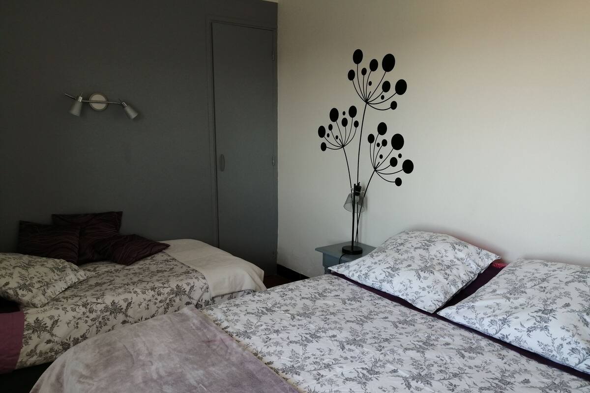 A bedroom with two beds, a nightstand, a wall-mounted lamp, and a door.