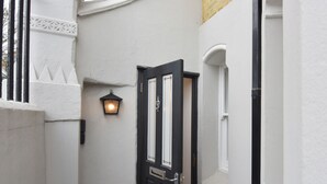 Ein schmaler Flur mit einer schwarzen Tür, einem kleinen Fenster und einer Wandleuchte.