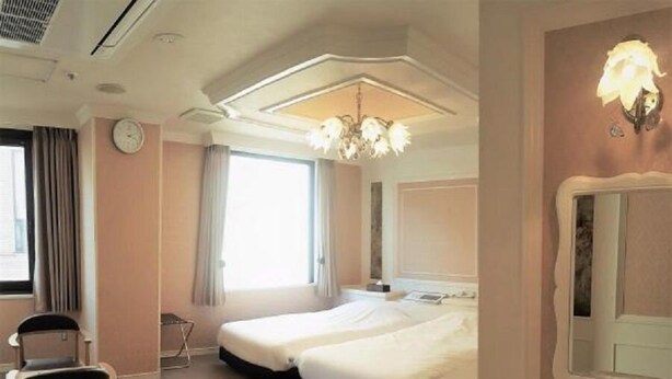 A hotel room with two beds, a desk, a chair, a chandelier, a clock, and a window with curtains. - Royal Hotel Shinokubo (Tokyo)
