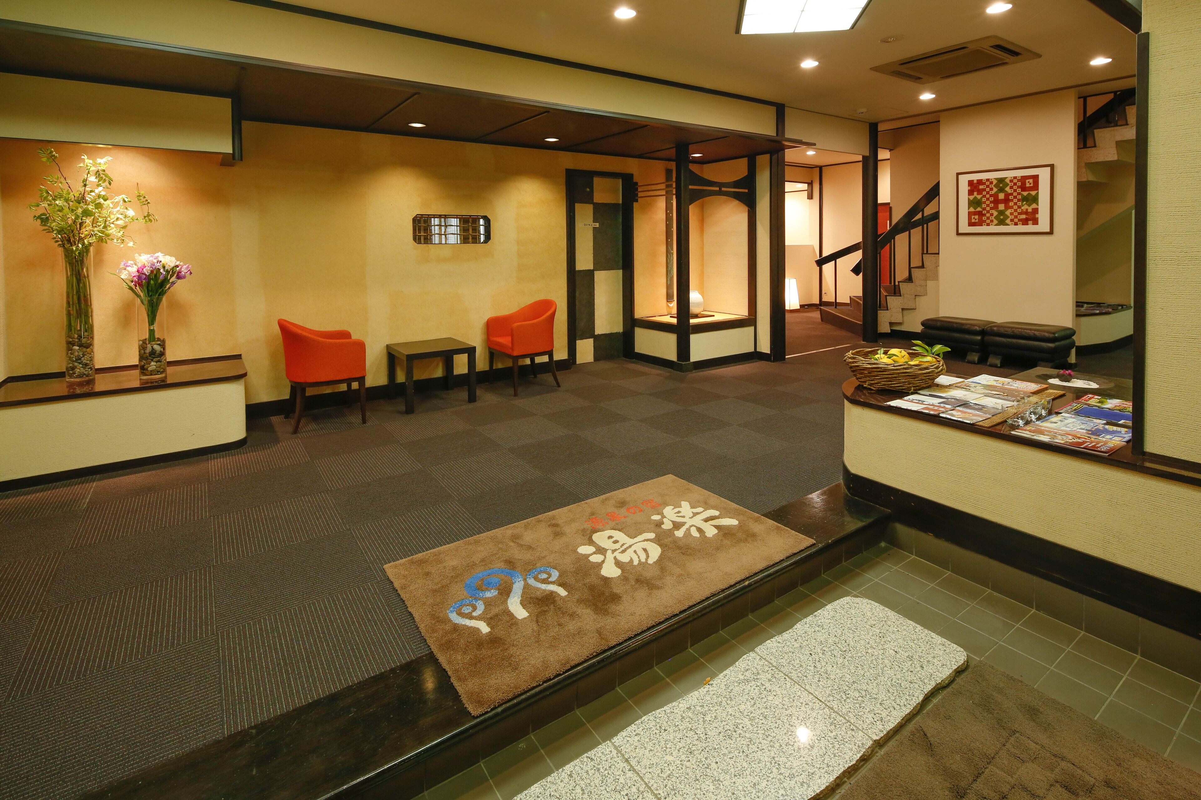 A hotel reception area with a reception desk, two orange chairs, a brown mat with Japanese characters, and a staircase in the background.