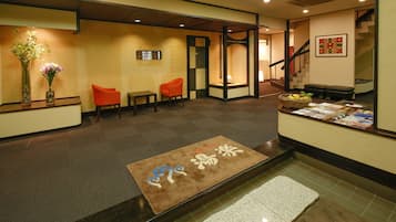 A hotel reception area with a reception desk, two orange chairs, a brown mat with Japanese characters, and a staircase in the background.
