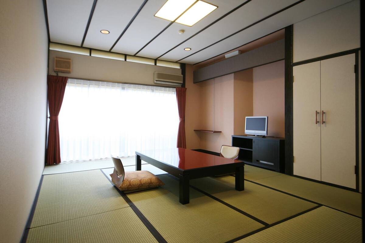 Ein traditioneller japanischer Raum mit Tatami-Boden, einem niedrigen Tisch, Kissen, einem Fernseher auf einer Anrichte und Schiebetüren.