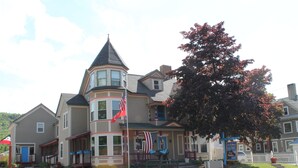 A two-story building with a prominent tower and bay windows, a flagpole with the American flag, and a large tree in front.