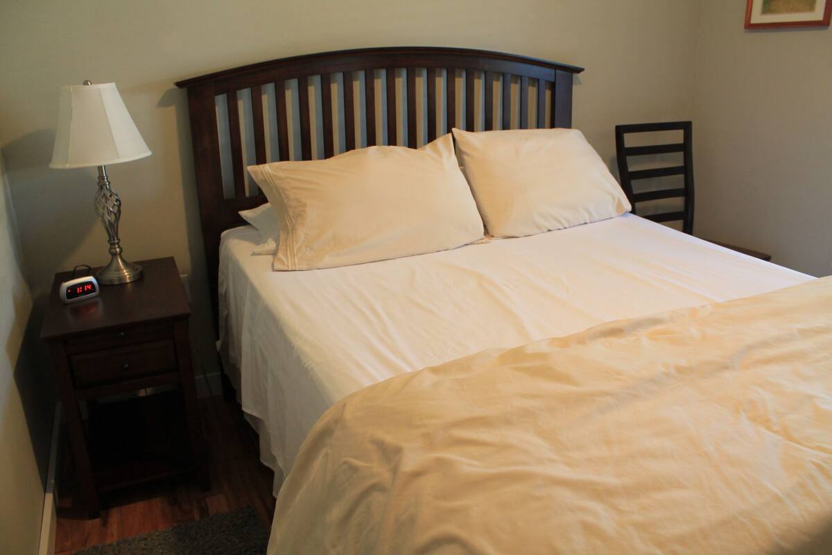 A neatly made bed with white linens, a wooden headboard, and a bedside table with a lamp and a clock.