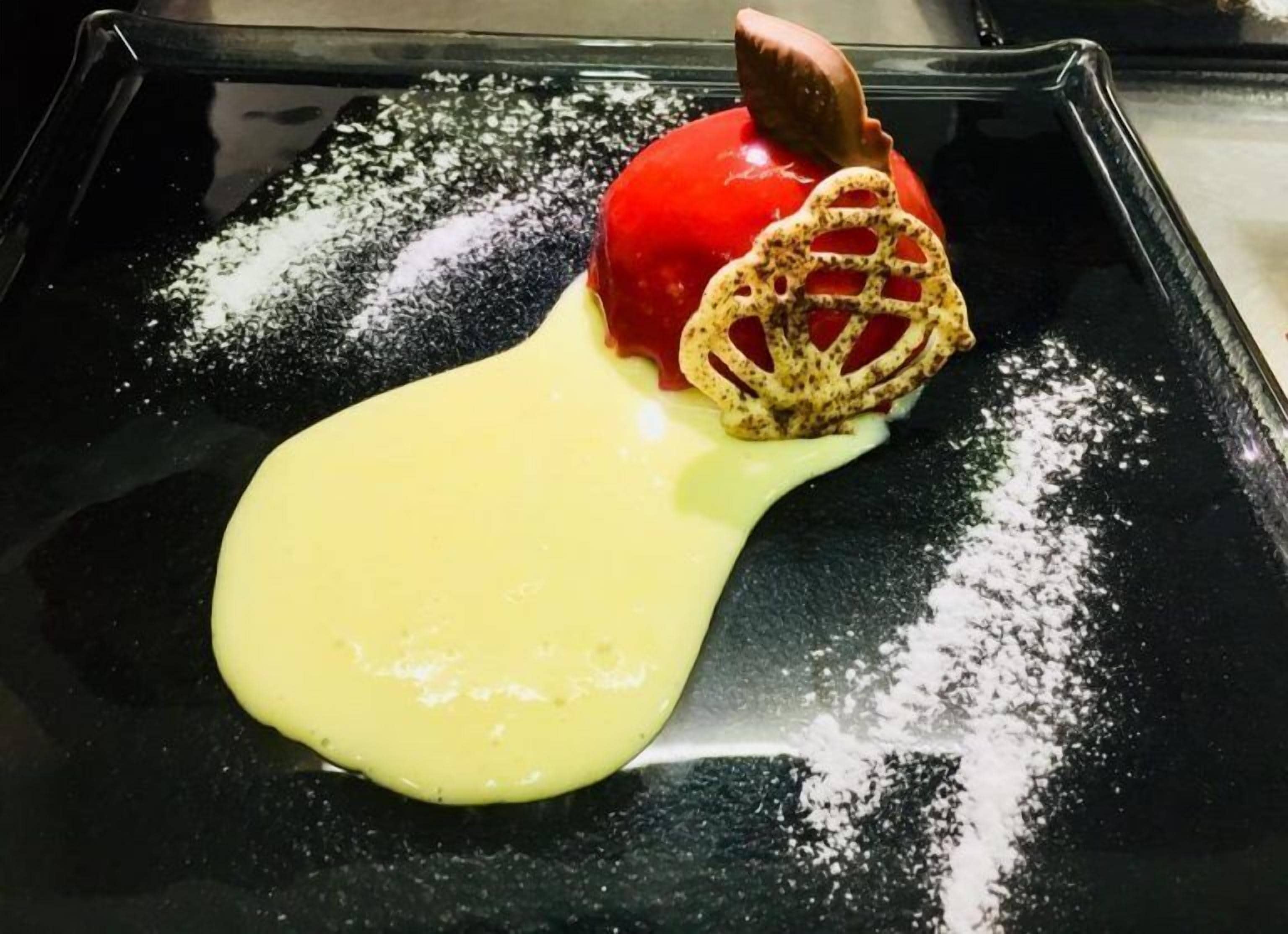 Postre en un plato negro con una fruta roja, una porción cubierta con rejilla y una salsa amarilla.