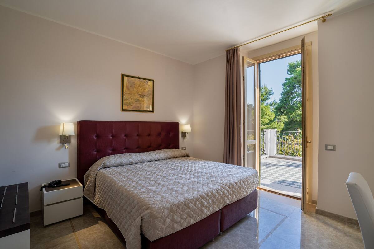 Un dormitorio con una cama grande, una mesita de noche, una lámpara y vistas al balcón exterior.