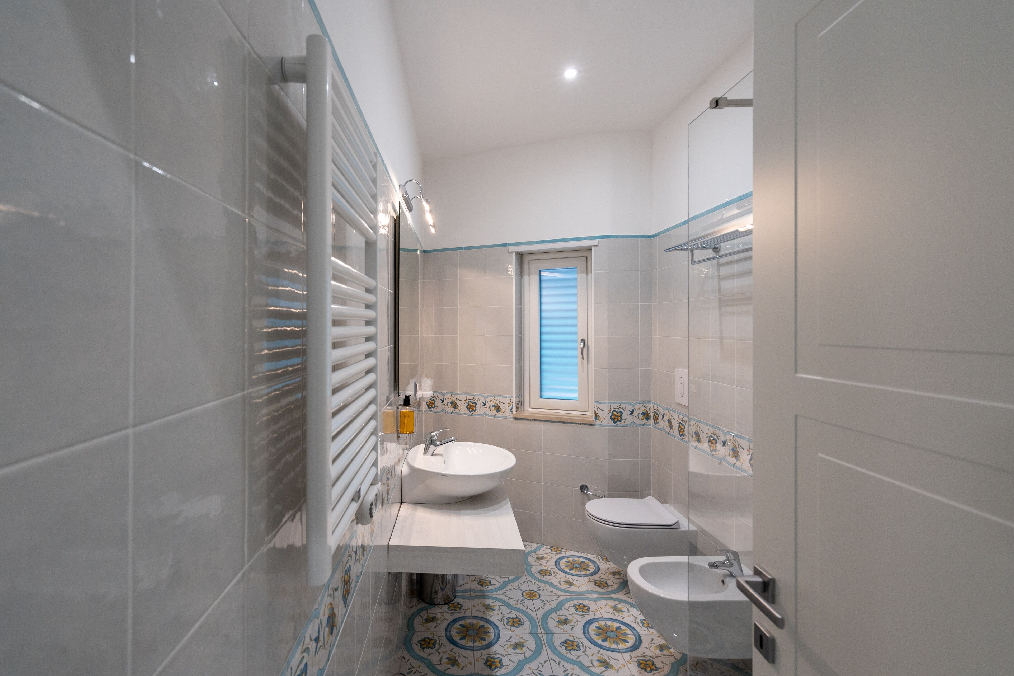 Ein modernes Badezimmer mit einem großen Waschbecken, einer Toilette und einem Bidet. Es verfügt über einen gefliesten Boden mit einem Dekor und ein Fenster mit blauen Rollläden.
