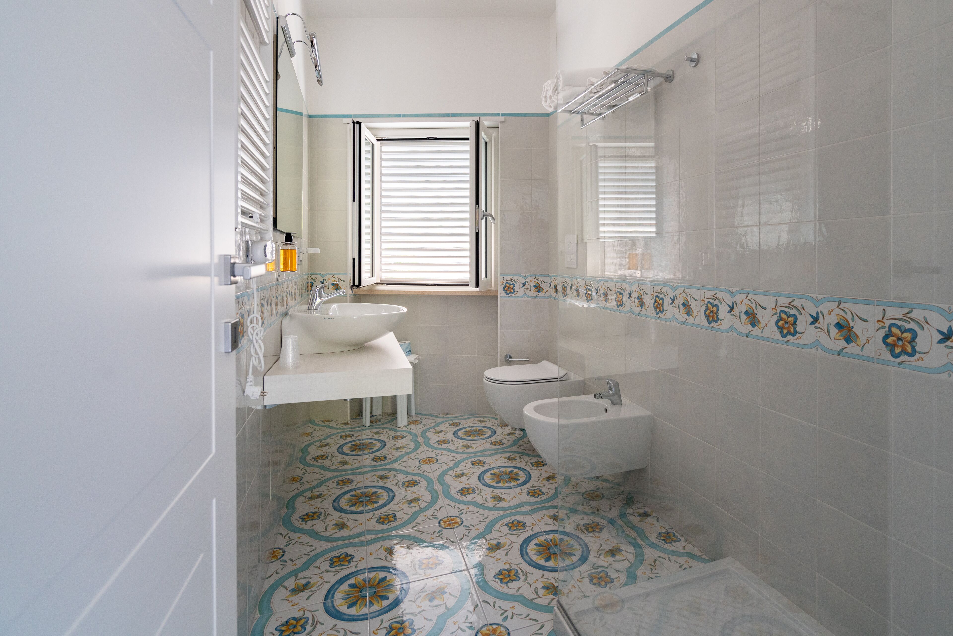 Un baño con lavamanos, inodoro y bidé, suelo de baldosas con estampado floral y una ventana con persianas.