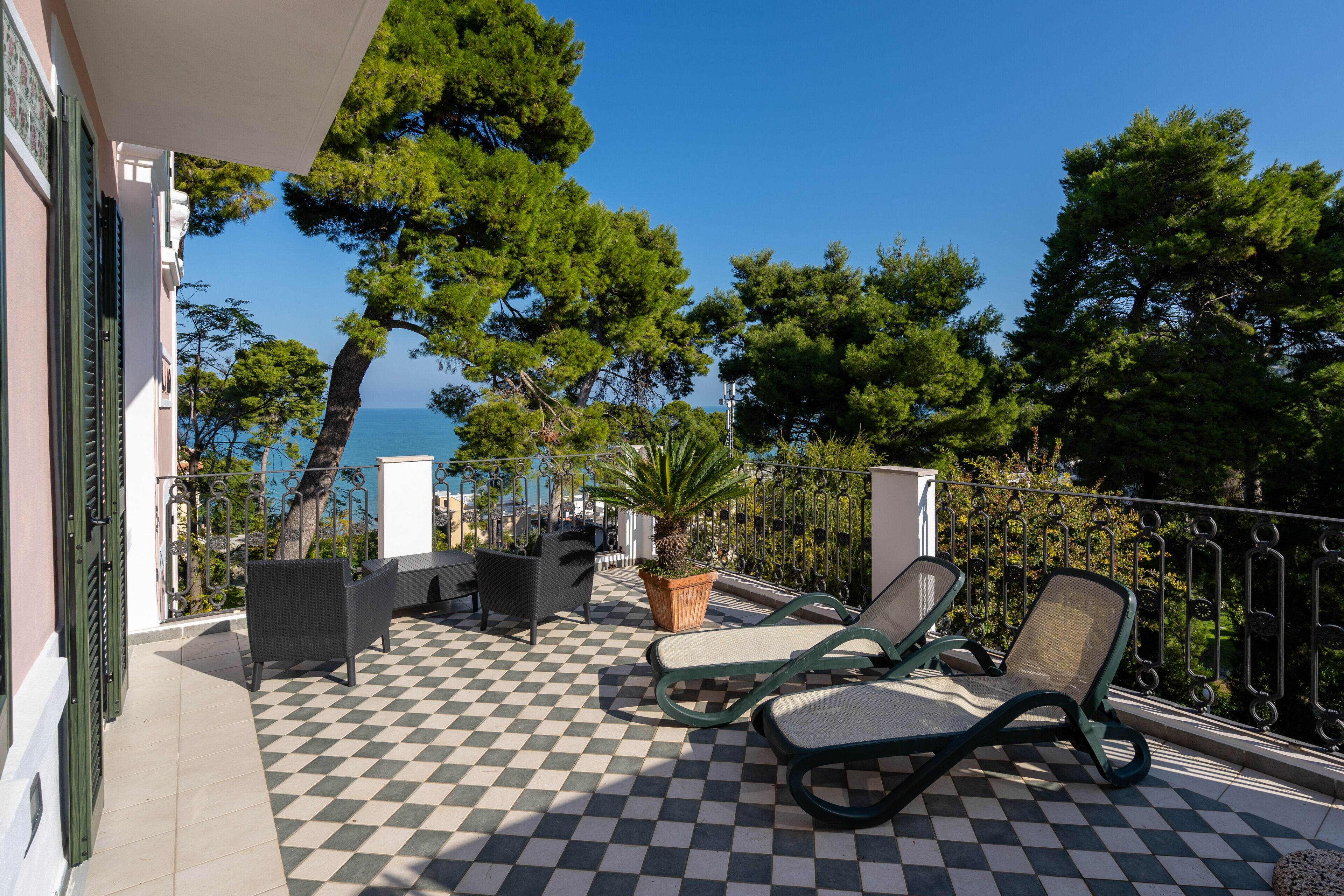 A balcony with checkered flooring, two lounge chairs, a potted plant, and a view of the sea and trees.