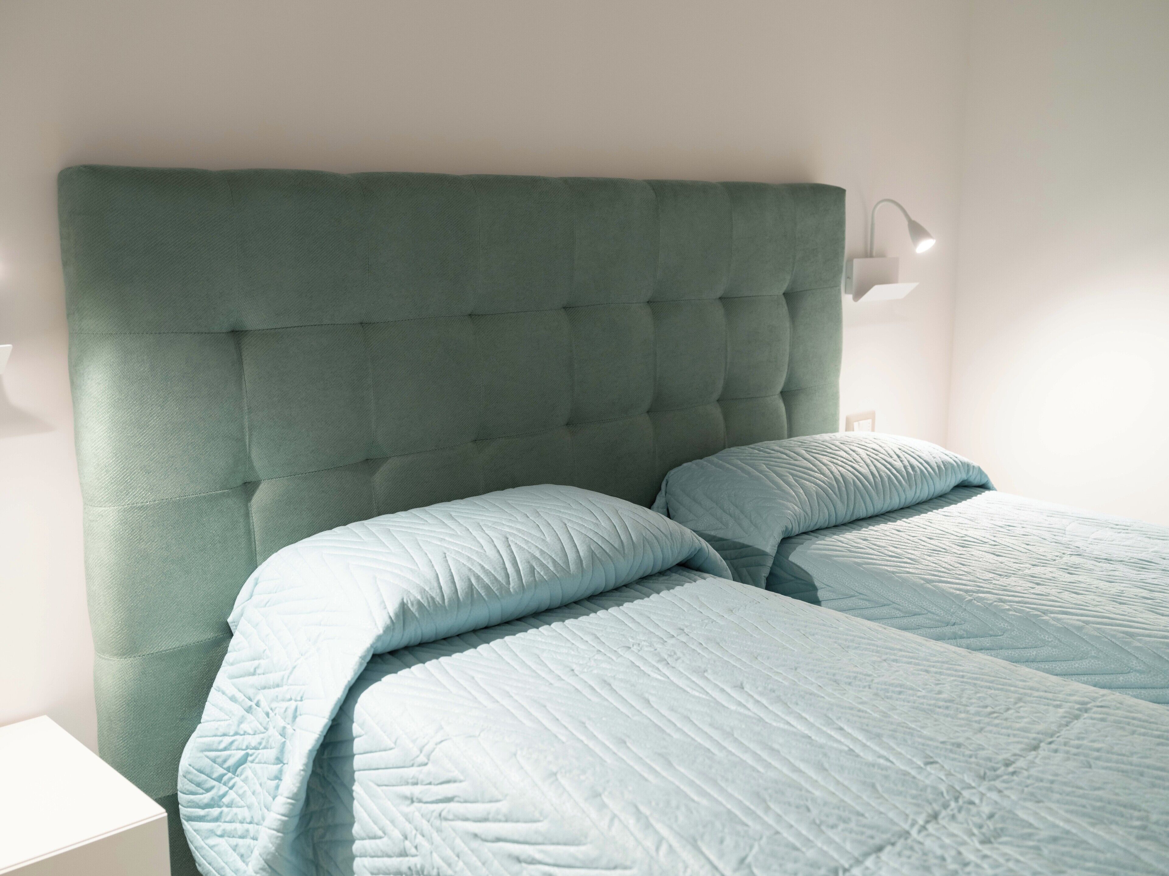 Un dormitorio con cabecera capitonada, dos camas con ropa de cama azul claro, una mesita de noche y una lámpara de pared.