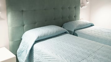 Un dormitorio con cabecera capitonada, dos camas con ropa de cama azul claro, una mesita de noche y una lámpara de pared.