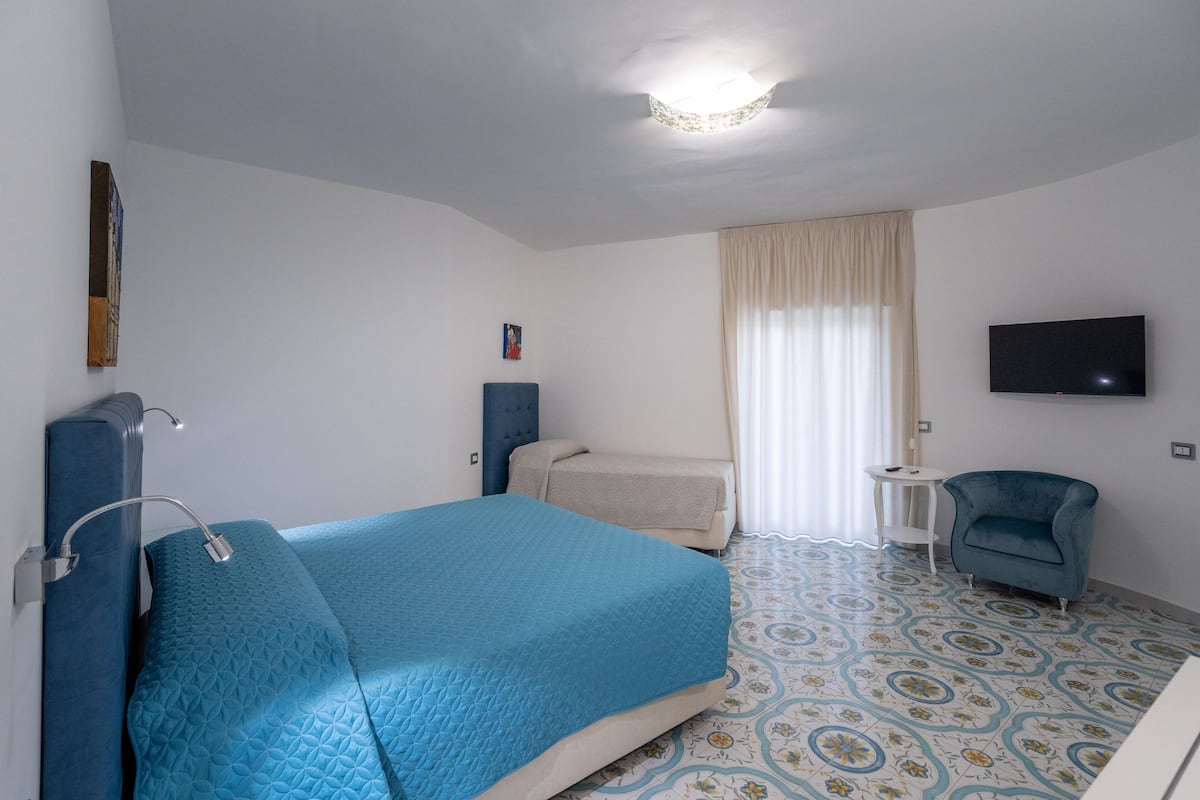 Una habitación de hotel con una cama, un sillón pequeño, un televisor y una ventana con cortinas.