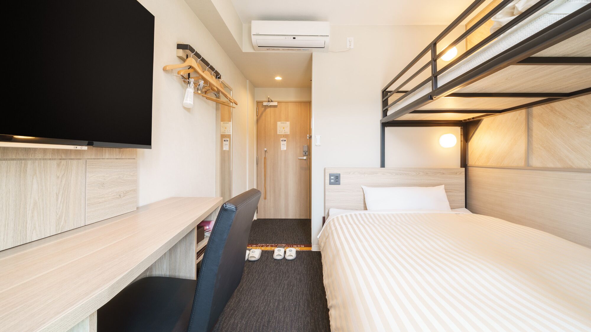 Une chambre d’hôtel avec un lit superposé, un bureau avec une chaise, une télévision et un porte-chaussures.