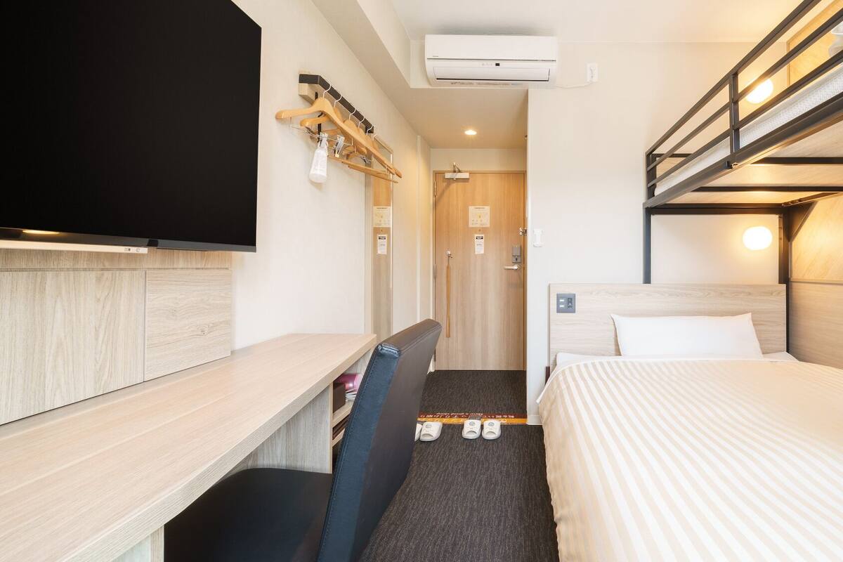 Una habitación de hotel con una cama litera, un escritorio con silla, un televisor y un zapatero.