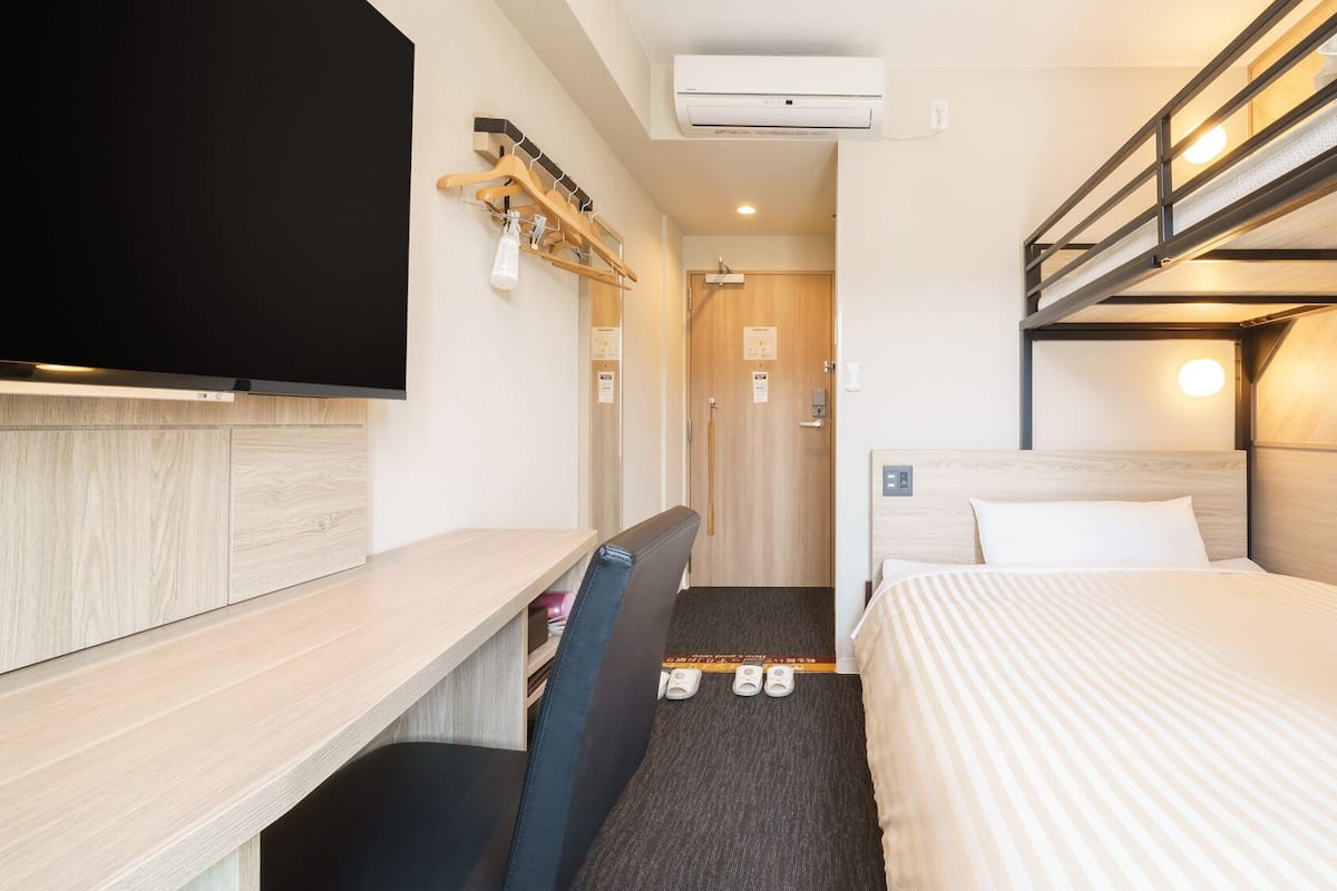 Une chambre d’hôtel avec un lit superposé, un bureau avec une chaise, une télévision et un porte-chaussures.