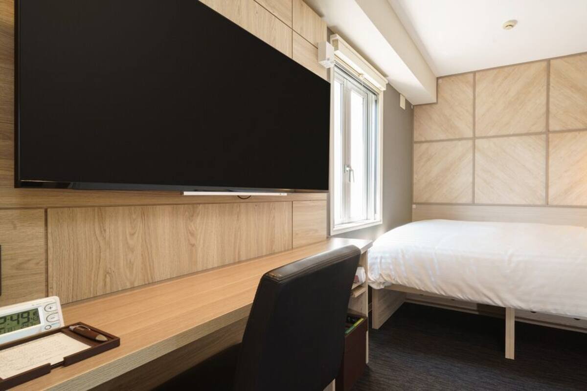 Habitación de hotel con cama, escritorio con silla, televisor de pantalla plana y ventana con persianas.
