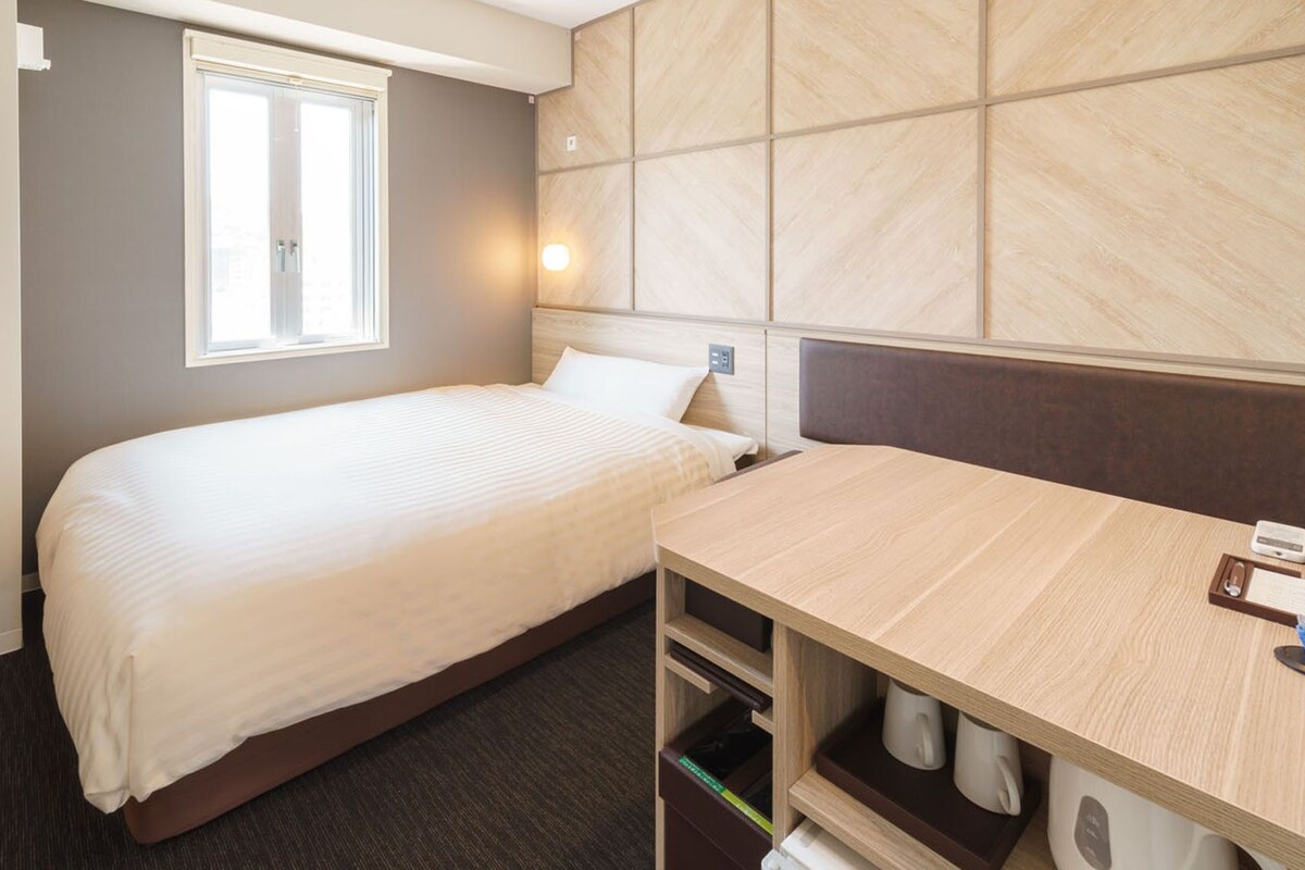 A hotel room with a bed, a desk, a window, and a wall-mounted light.