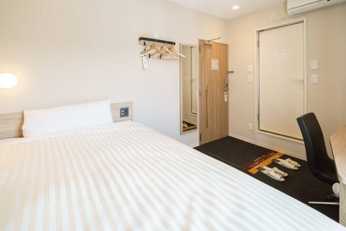 Una habitación de hotel con cama, una silla, perchero, armario y espejo.