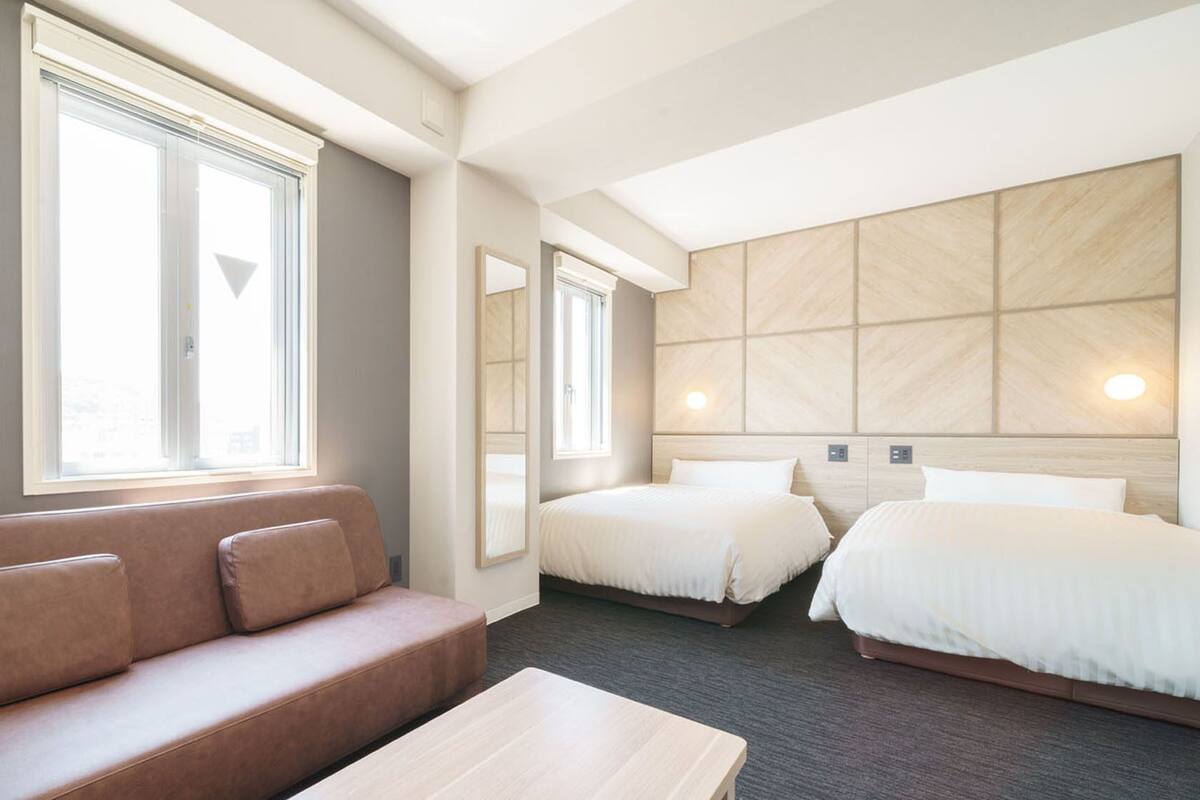 Habitación de hotel con sofá, dos camas, cabecero de madera y ventanas grandes.