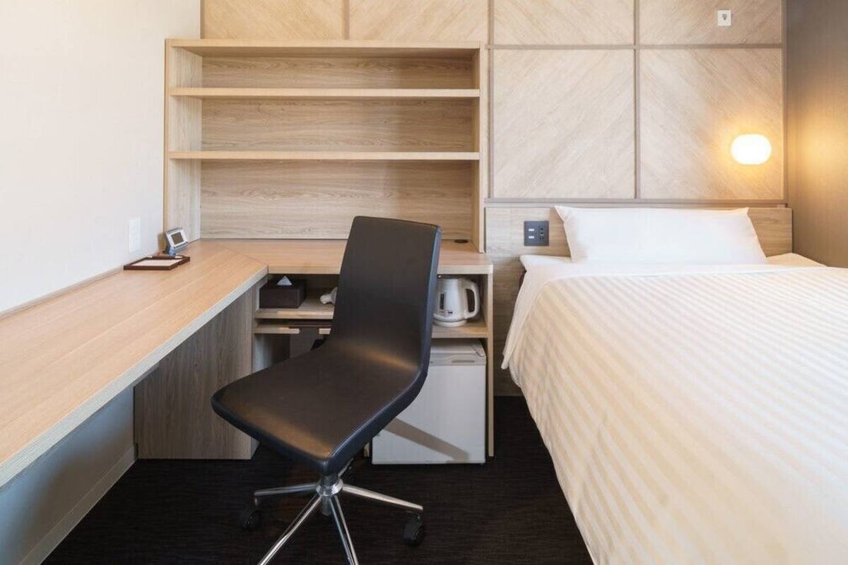 Habitación de hotel con cama, escritorio con silla, kitchenette pequeño y estante de pared.