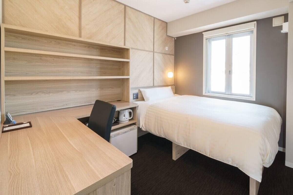 Un dormitorio con cama, escritorio, silla, mesita de noche pequeña y ventana.