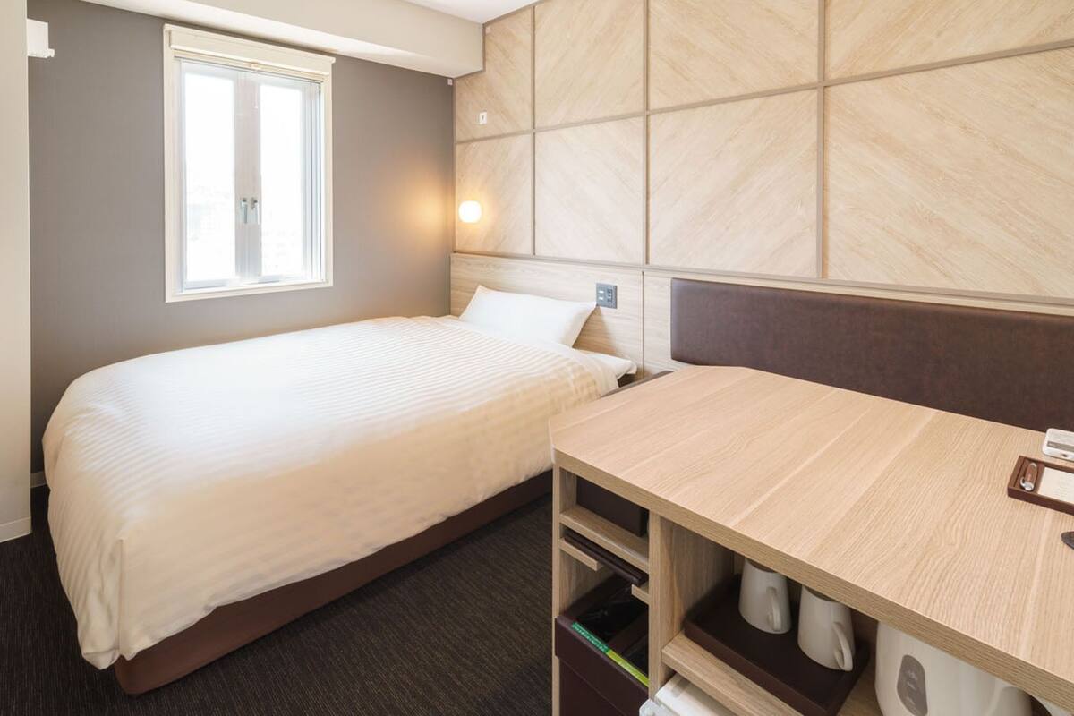Habitación de hotel con cama, escritorio, ventana y lámpara fijada a la pared.