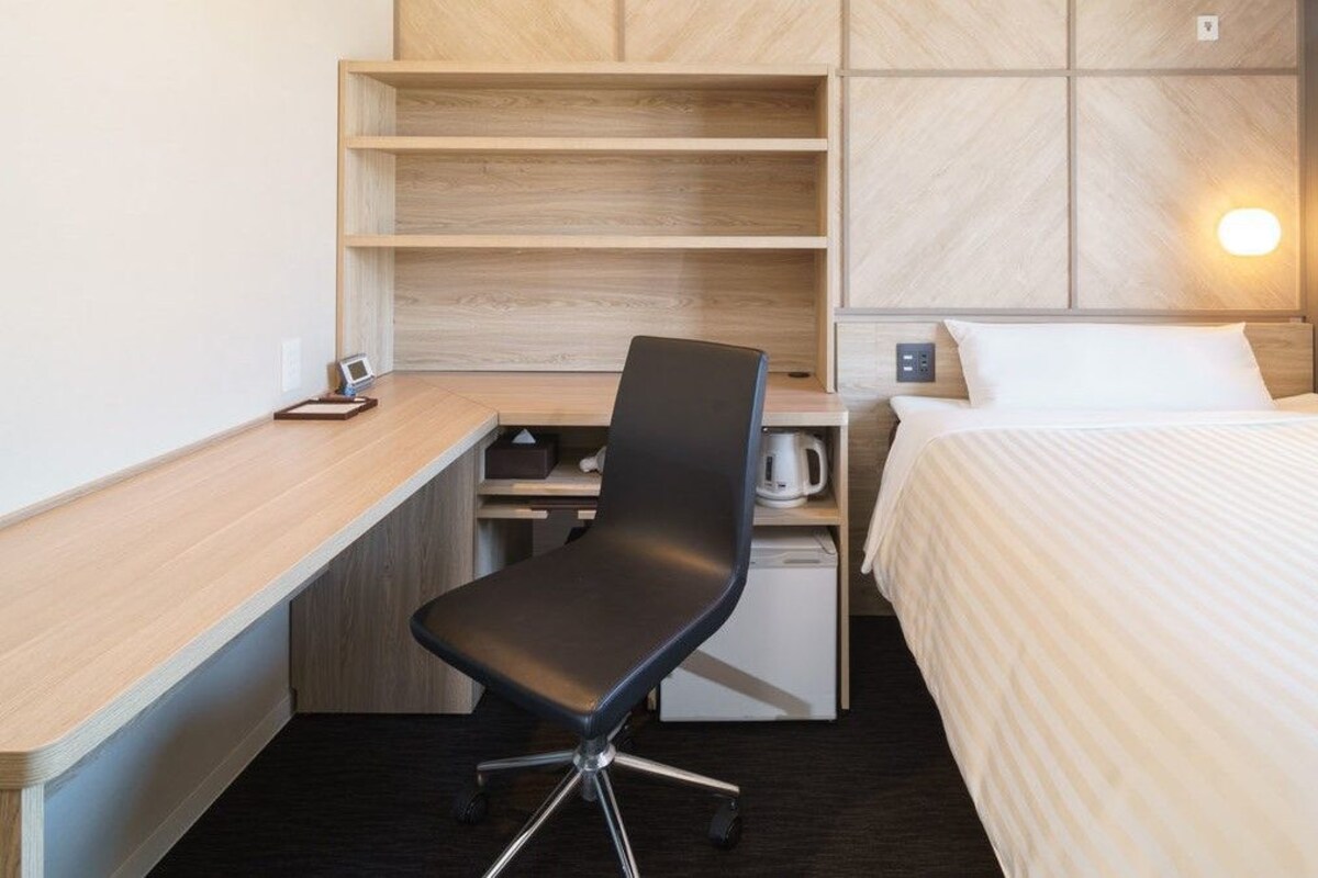 A hotel room with a bed, a desk with a chair, a small kitchenette, and a wall-mounted shelf.