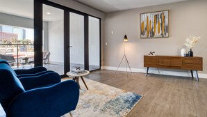 Uma sala de estar moderna com uma grande porta de vidro deslizante para uma varanda, um sofá azul, uma pequena mesa redonda com livros, um abajur e um aparador de madeira com objetos decorativos.