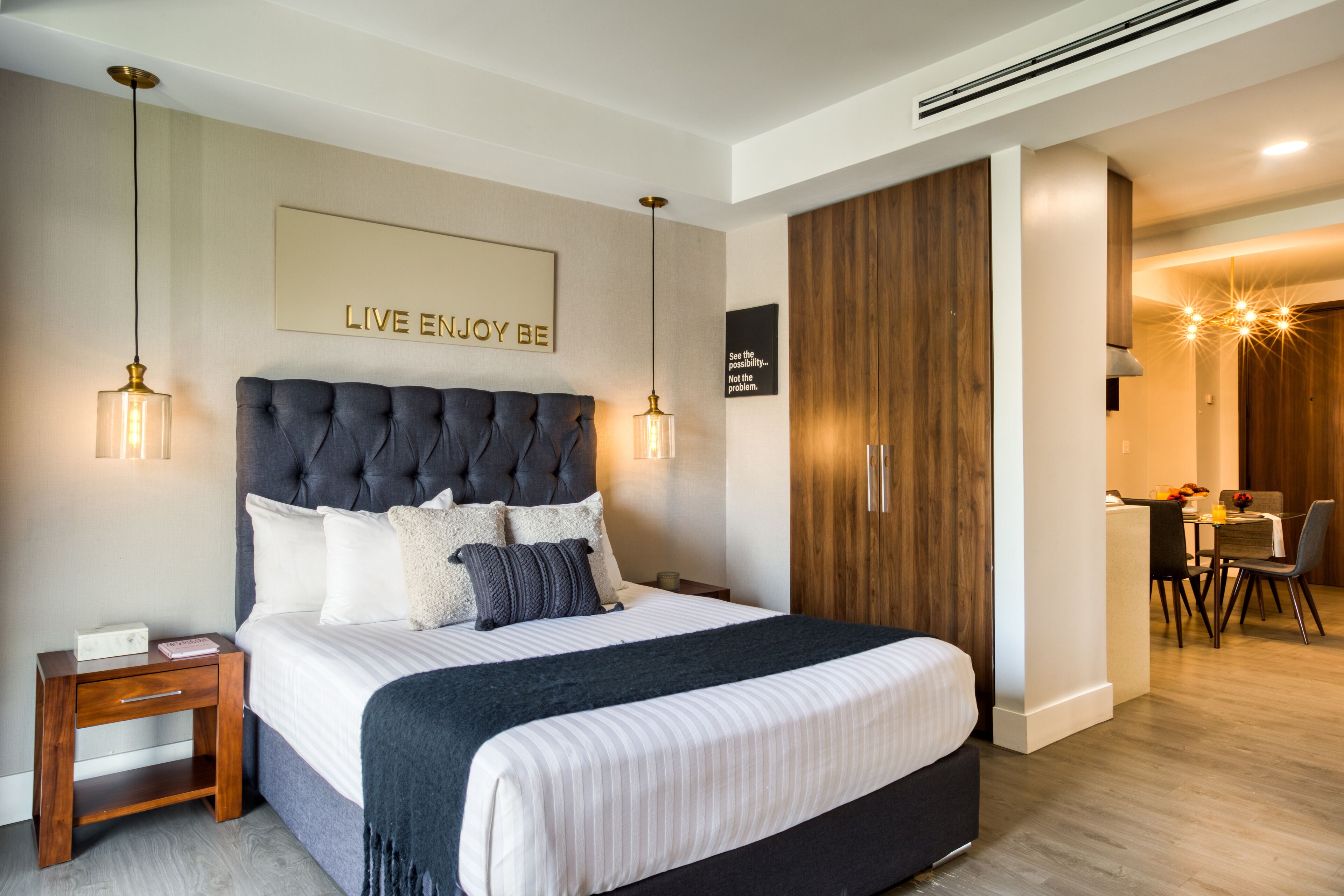 A modern bedroom with a large bed, bedside tables, a wooden wardrobe, and a dining area visible through an open door.