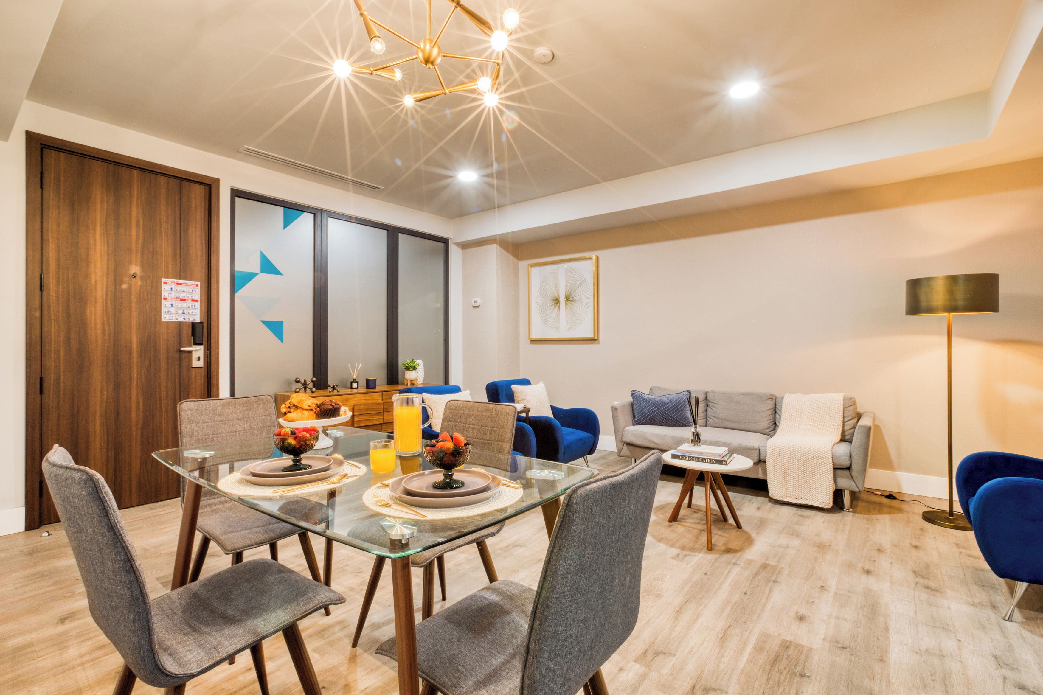 A modern dining area with a glass table, grey upholstered chairs, and a blue sofa in the background.