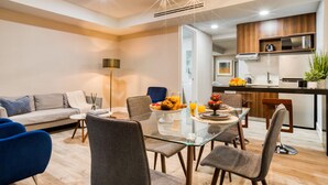 A modern living room with a glass dining table, grey and blue chairs, a sofa, and a kitchen in the background.