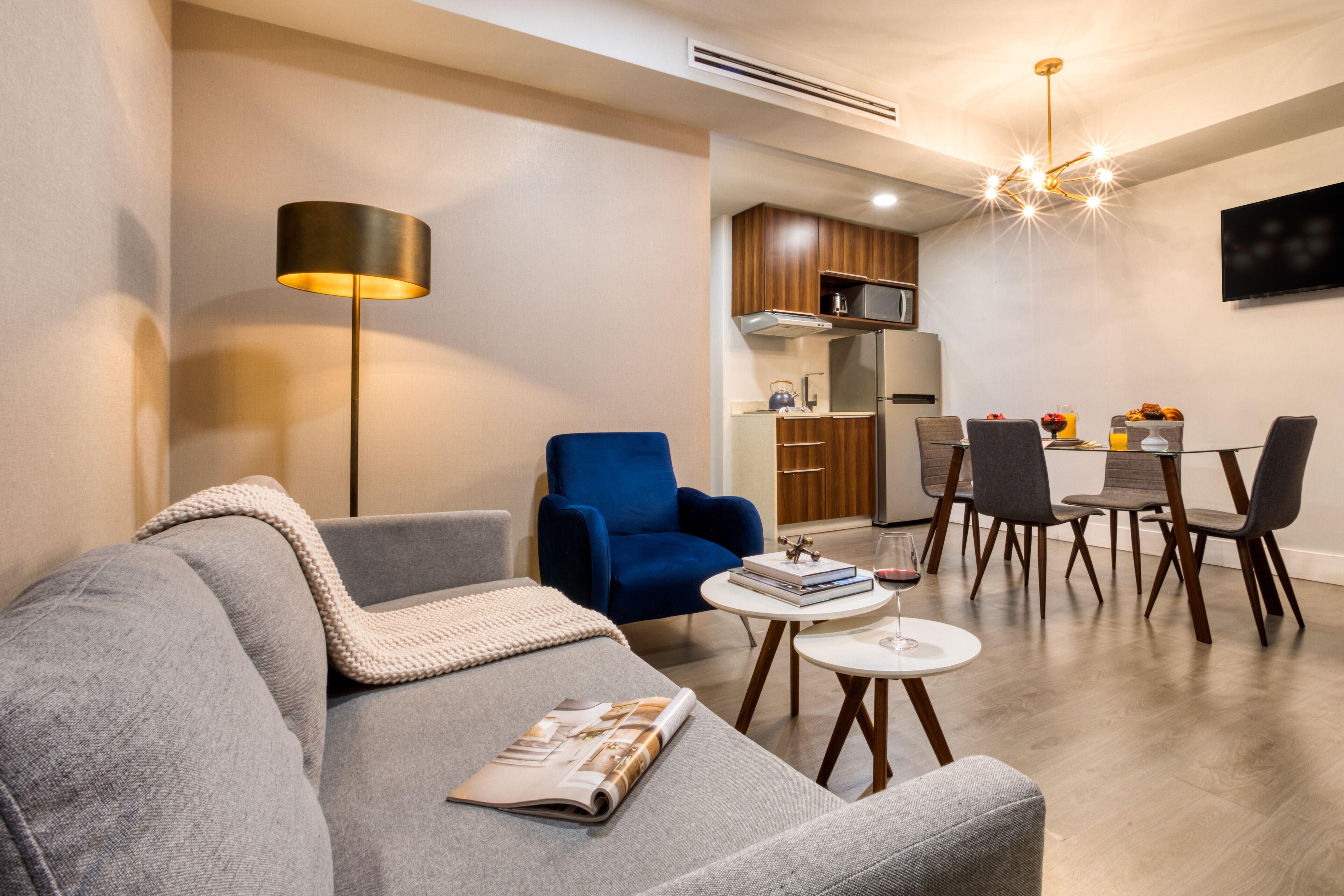 A modern living room with a grey sofa, a blue armchair, and a small round table with a magazine. A dining area with a table and chairs is visible in the background.