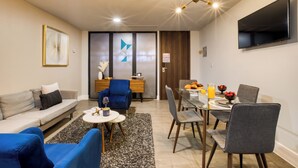 Uma sala de estar moderna com um sofá, poltronas azuis, uma mesa de jantar de vidro com cadeiras, uma TV fixada na parede e um lustre.