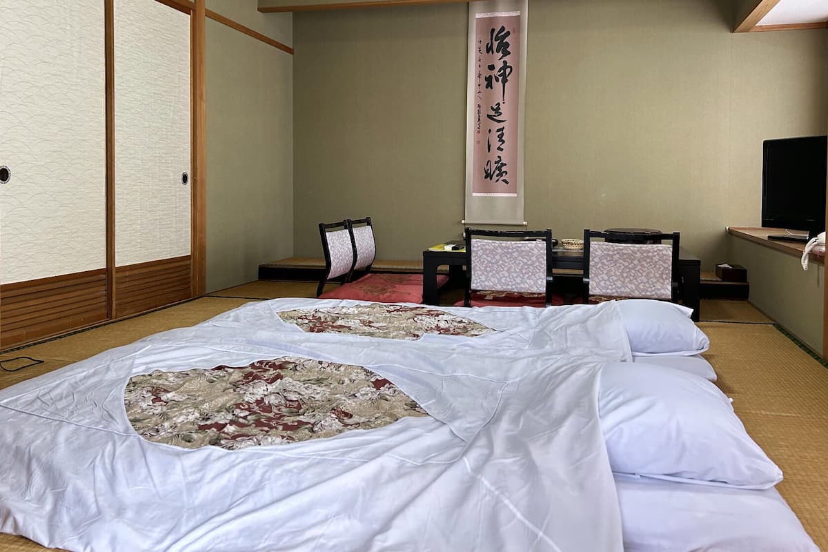 Una habitación tradicional japonesa con suelo de tatami, futón, pantalla shoji, escritorio con teléfono y televisión.
