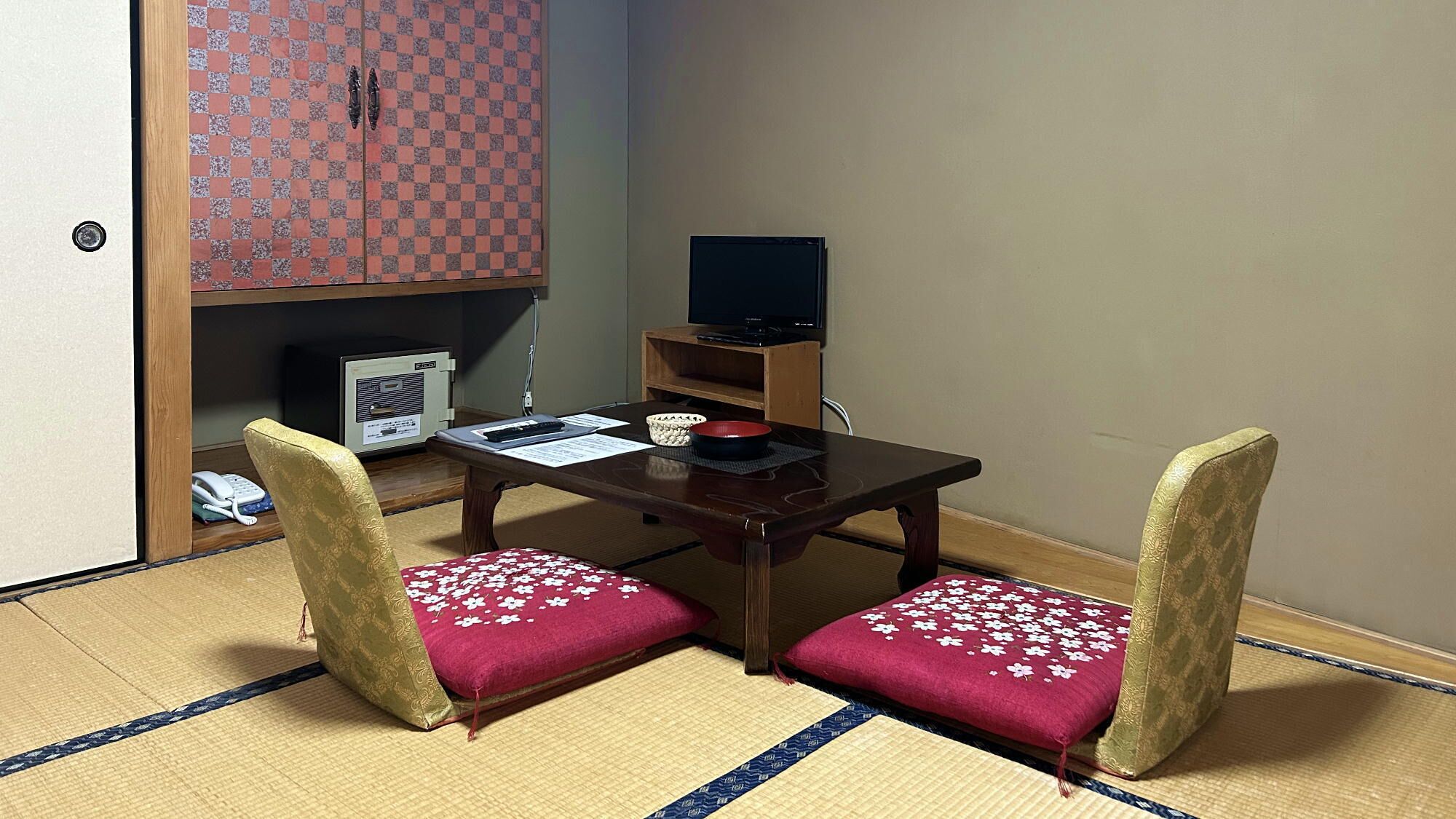 Una habitación japonesa tradicional con suelo de tatami, una mesa baja, dos sillas con cojines florales, un televisor y un armario con estampado de cuadros.