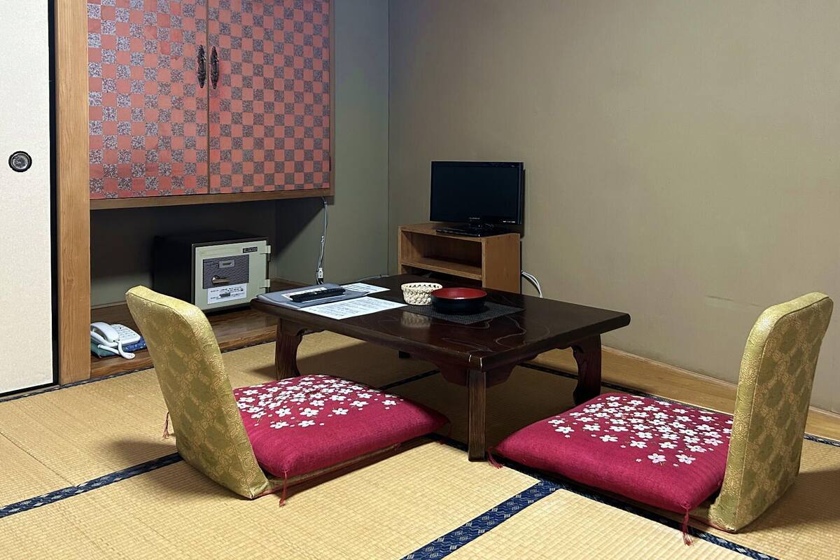 Une pièce de style japonais traditionnel, avec un sol recouvert de tatamis, une table basse, deux chaises dotées de coussins à motifs floraux, un téléviseur et un meuble au motif à carreaux.