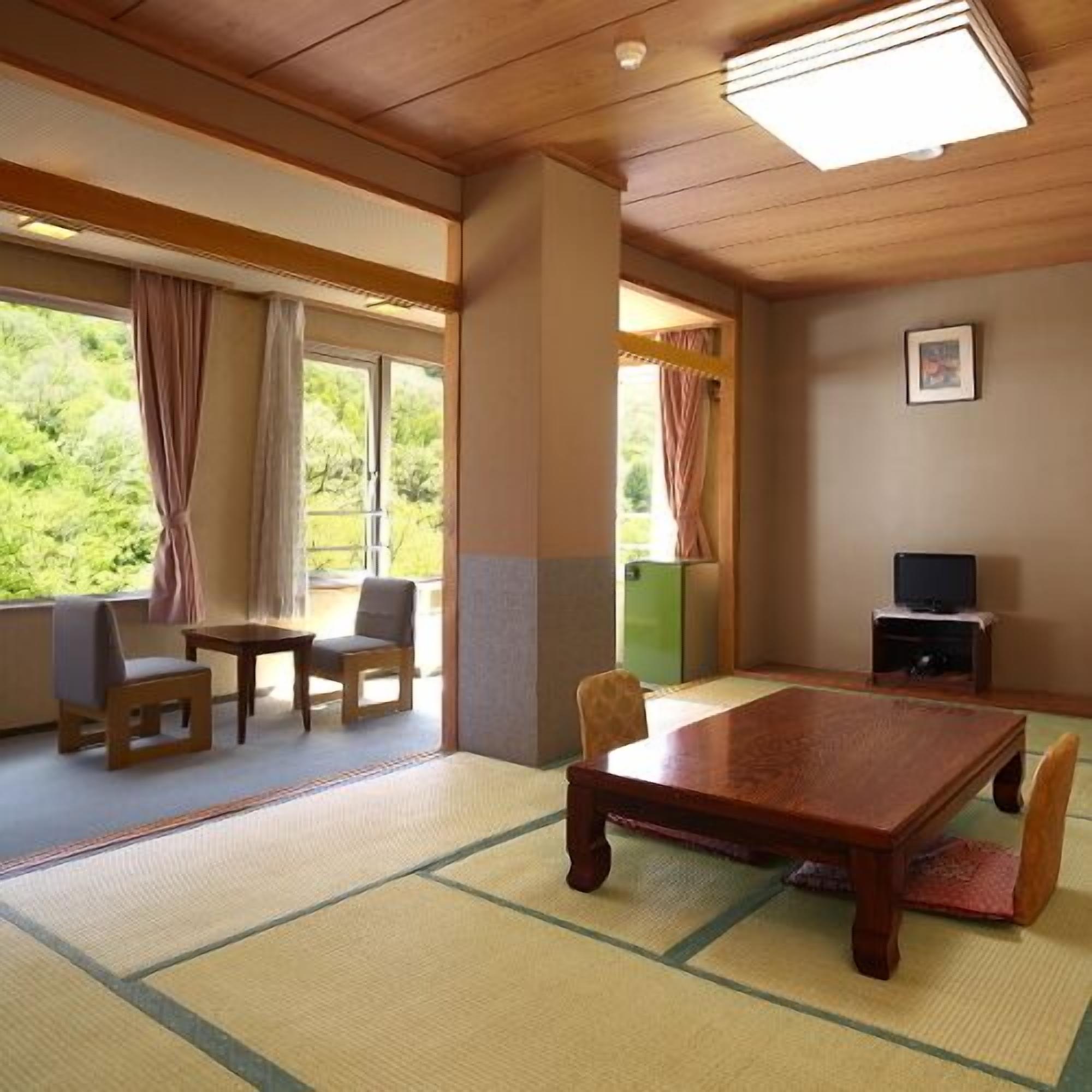Una habitación con esteras de tatami, una mesa de madera, sillas, un televisor y una ventana con cortinas.
