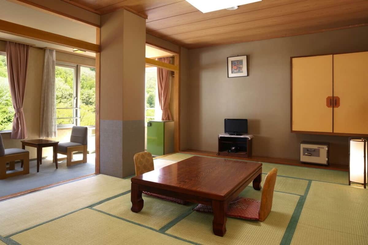 Une pièce avec un sol recouvert de tatamis, une table en bois, des chaises, un téléviseur et un meuble.
