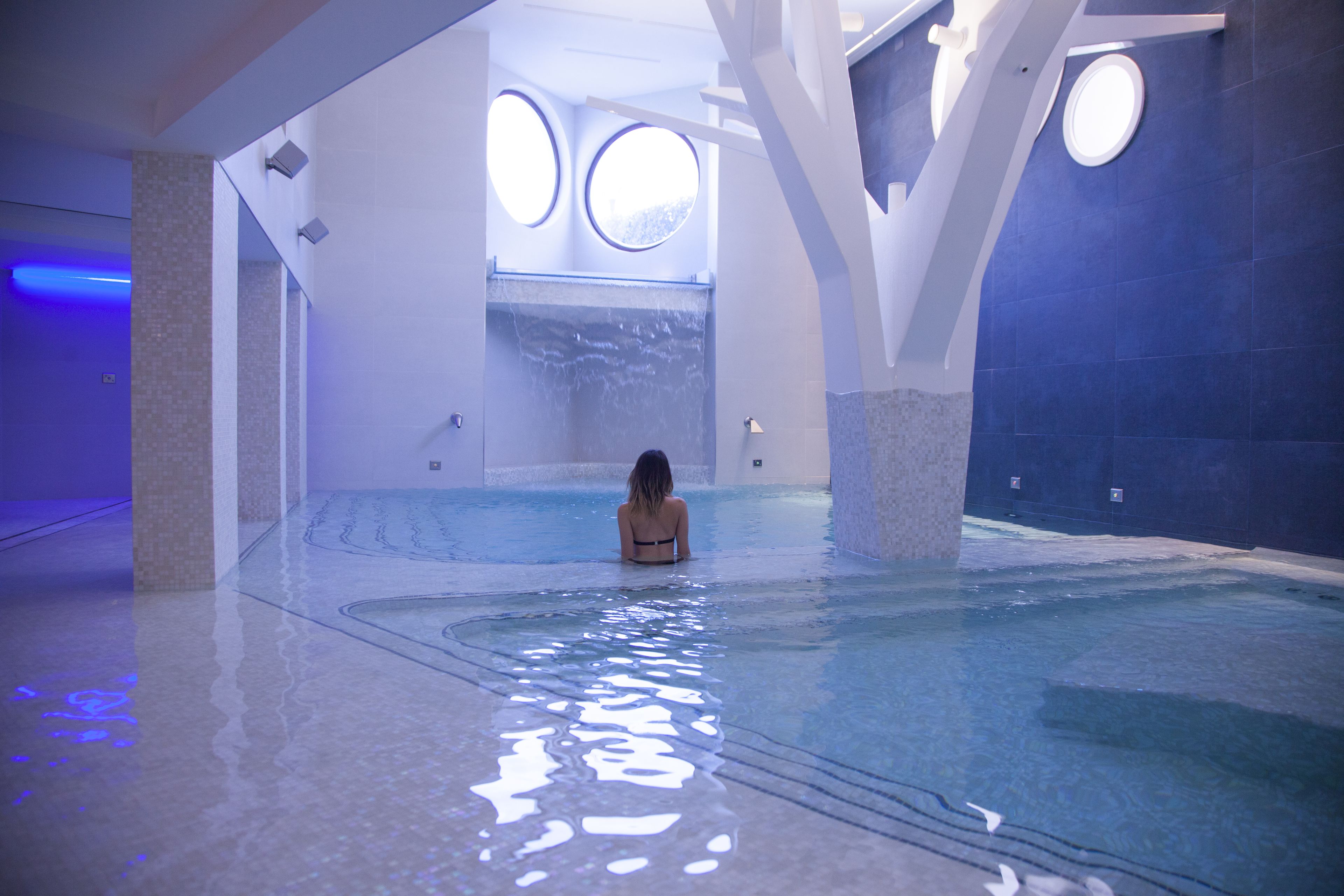 A woman sitting in a large indoor swimming pool with a modern architectural design, featuring large windows and a clean, reflective floor.