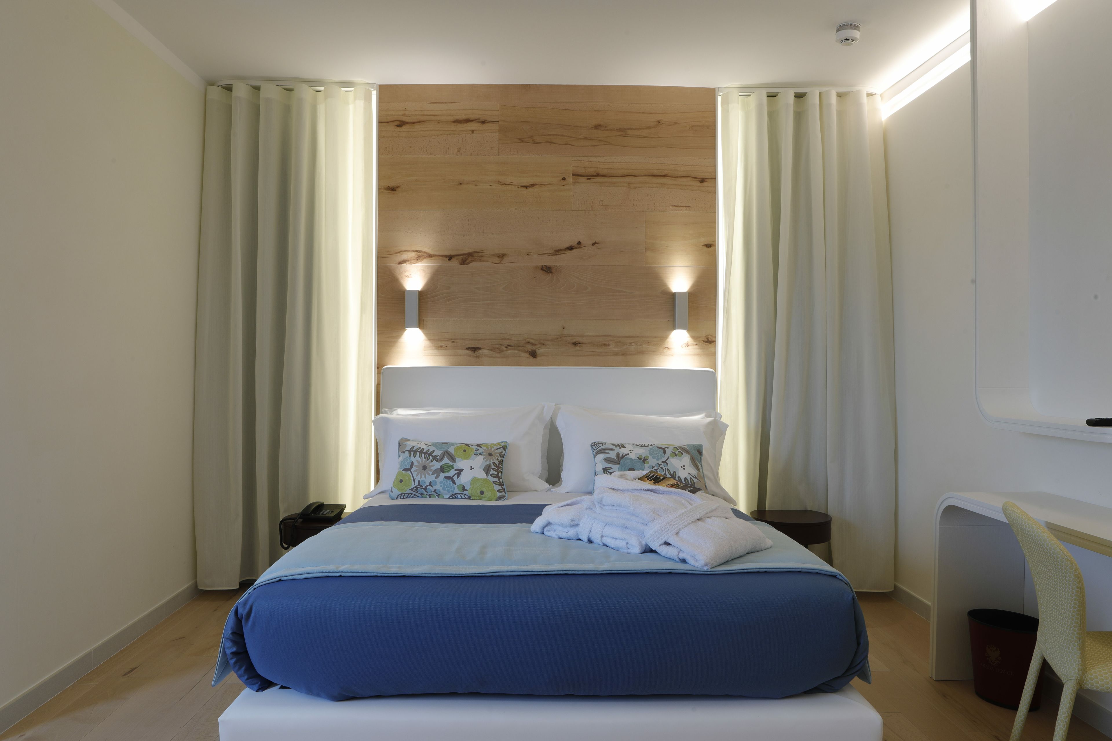 Uma cama bem arrumada com colcha azul e almofadas brancas, cabeceira de madeira e uma pequena mesa de cabeceira com abajur.