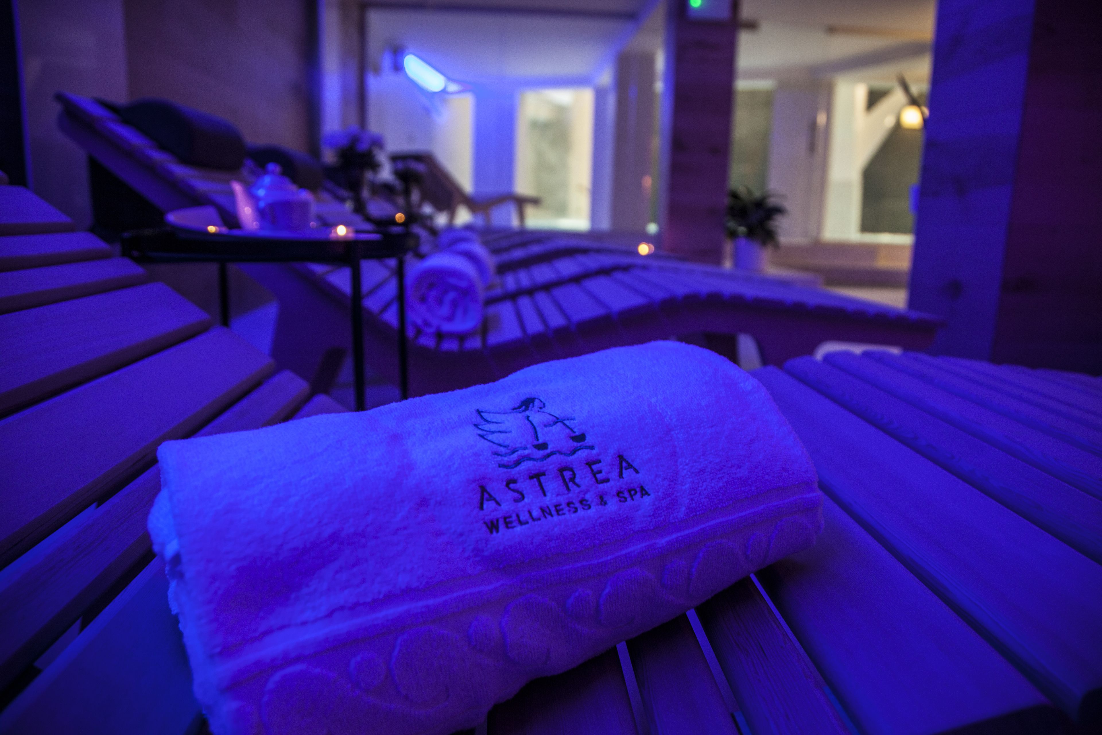 Een handdoek met de branding 'ASTREA WELLNESS & SPA' op een houten bankje in een kamer met weinig licht en uitzicht op een modern interieur.