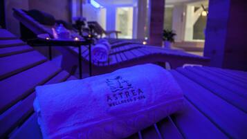 Een handdoek met de branding 'ASTREA WELLNESS & SPA' op een houten bankje in een kamer met weinig licht en uitzicht op een modern interieur.