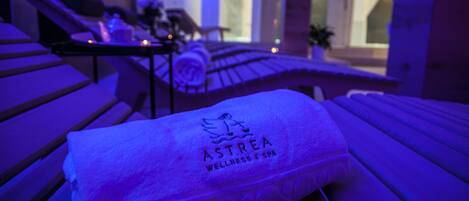 Una toalla con la marca "ASTREA WELLNESS & SPA" sobre un banco de madera en una habitación tenuemente iluminada con vistas a un interior moderno.