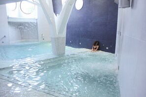 A large indoor swimming pool with a person in the water, white pillars, and a blue tiled wall. - Astrea Wellness & Spa (Altavilla Silentina)